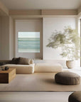 A serene and minimalist living room with a large window, a white sofa, armchairs, and a low coffee table. The wall features a print of a calm beach scene.