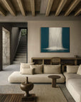 A painting with abstract blue lines displayed above a beige sofa in a room with wooden beams and a stone staircase in the background.