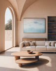 A minimalist designed living room with a large framed artwork above the sofa, a circular wooden coffee table in the foreground, and a large arched door in the background leading to another room or balcony.