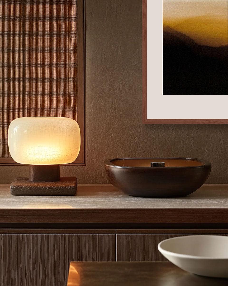 A framed artwork titled 'Rising Lines' hanging on a wall, with a illuminated table lamp and a bowl on a shelf below it.