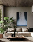 A modern living room with a large abstract painting in shades of blue and white, surrounded by floor-to-ceiling windows with a view of greenery outside. The room features a neutral color scheme with wooden elements and a cozy sofa setup.
