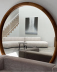 Round wooden-framed mirror reflecting a modern living room with a sofa and artwork.