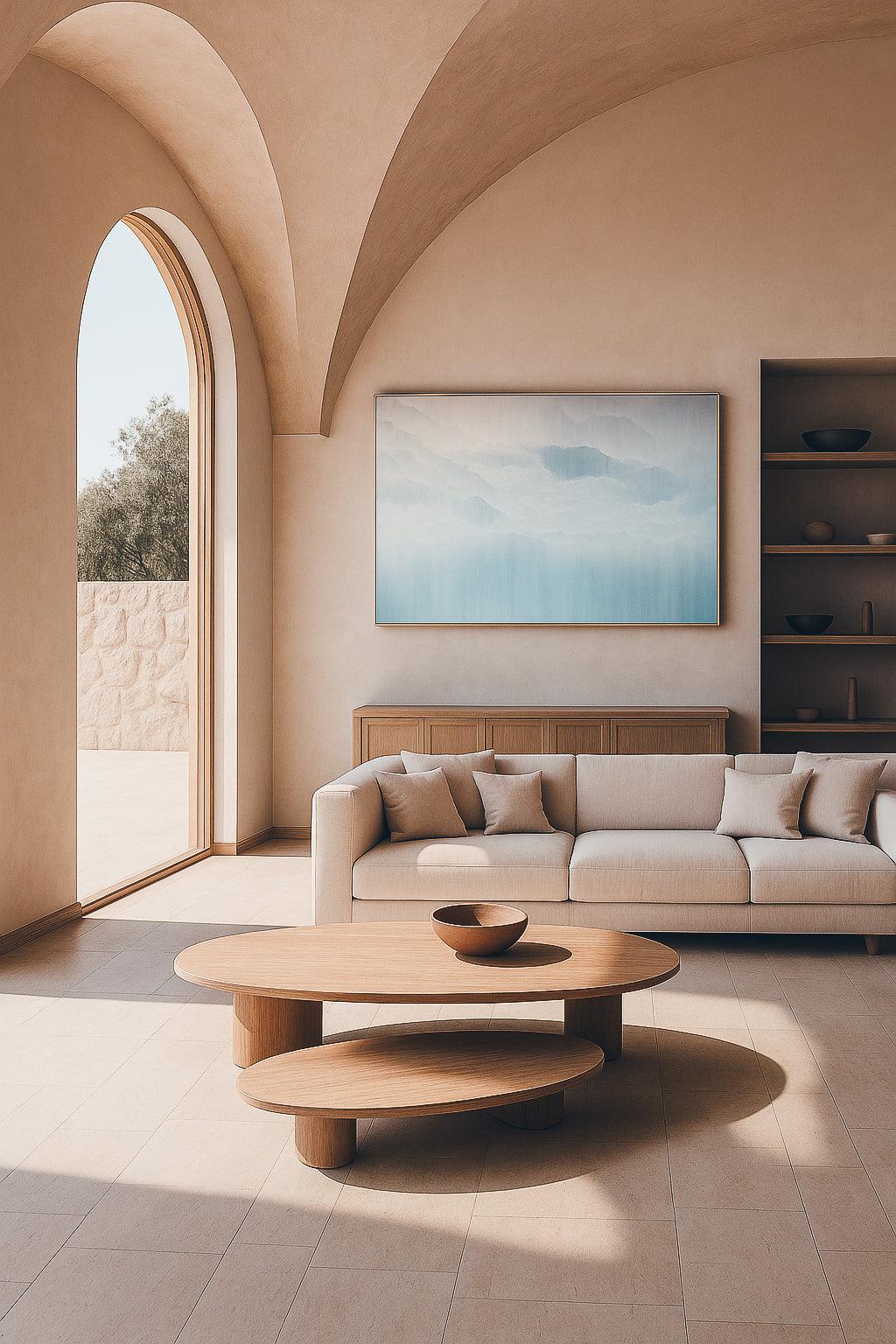 A minimalist designed living room with a large framed artwork above the sofa, a circular wooden coffee table in the foreground, and a large arched door in the background leading to another room or balcony.