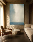 A large, minimalist artwork depicting a serene sky and sea scene, displayed on a wall in a well-lit living room setting with a sofa and armchair.
