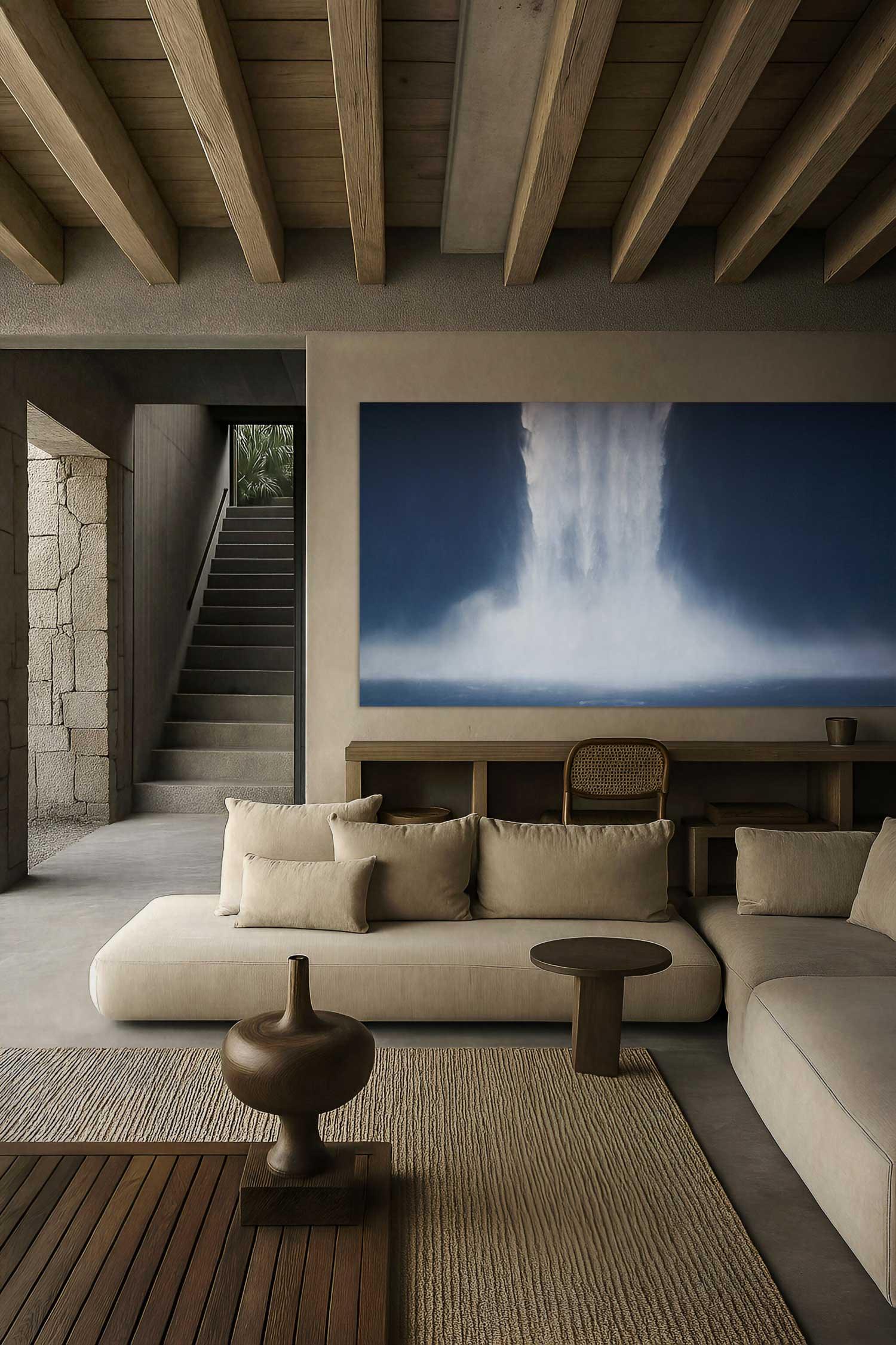 A spacious, well-lit room featuring a beige sofa in front of a large painting depicting a natural scene with waterfalls. The room has a wooden floor, visible beams on the ceiling, and a modern staircase in the background.