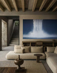 A spacious, well-lit room featuring a beige sofa in front of a large painting depicting a natural scene with waterfalls. The room has a wooden floor, visible beams on the ceiling, and a modern staircase in the background.