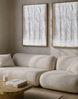 Two framed artworks depicting white tree-like figures against a brown background, displayed above a beige sofa in a well-lit room with large windows.