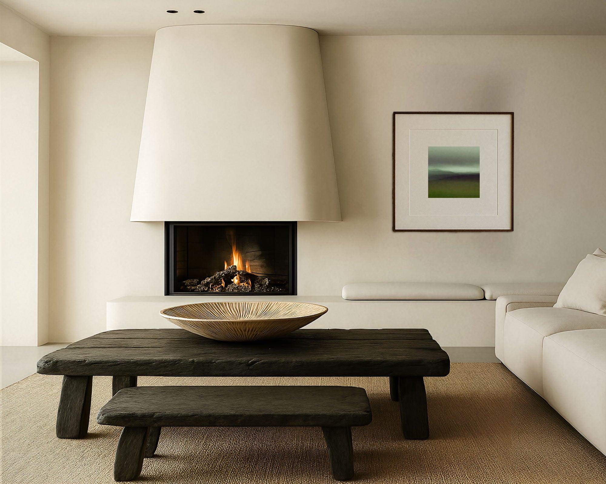 A framed artwork with a serene landscape depiction, hanging on the wall above a dark wooden bench and a low table in a minimalist living space with a fireplace in the background.
